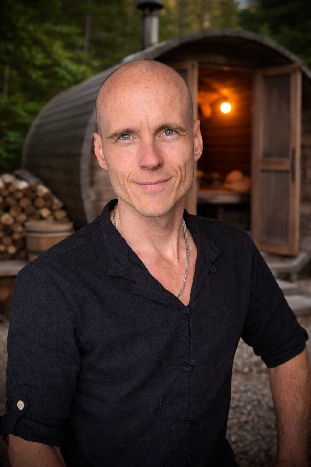 David Drimmel standing in front of a wooden sauna with a warm light inside, surrounded by trees.