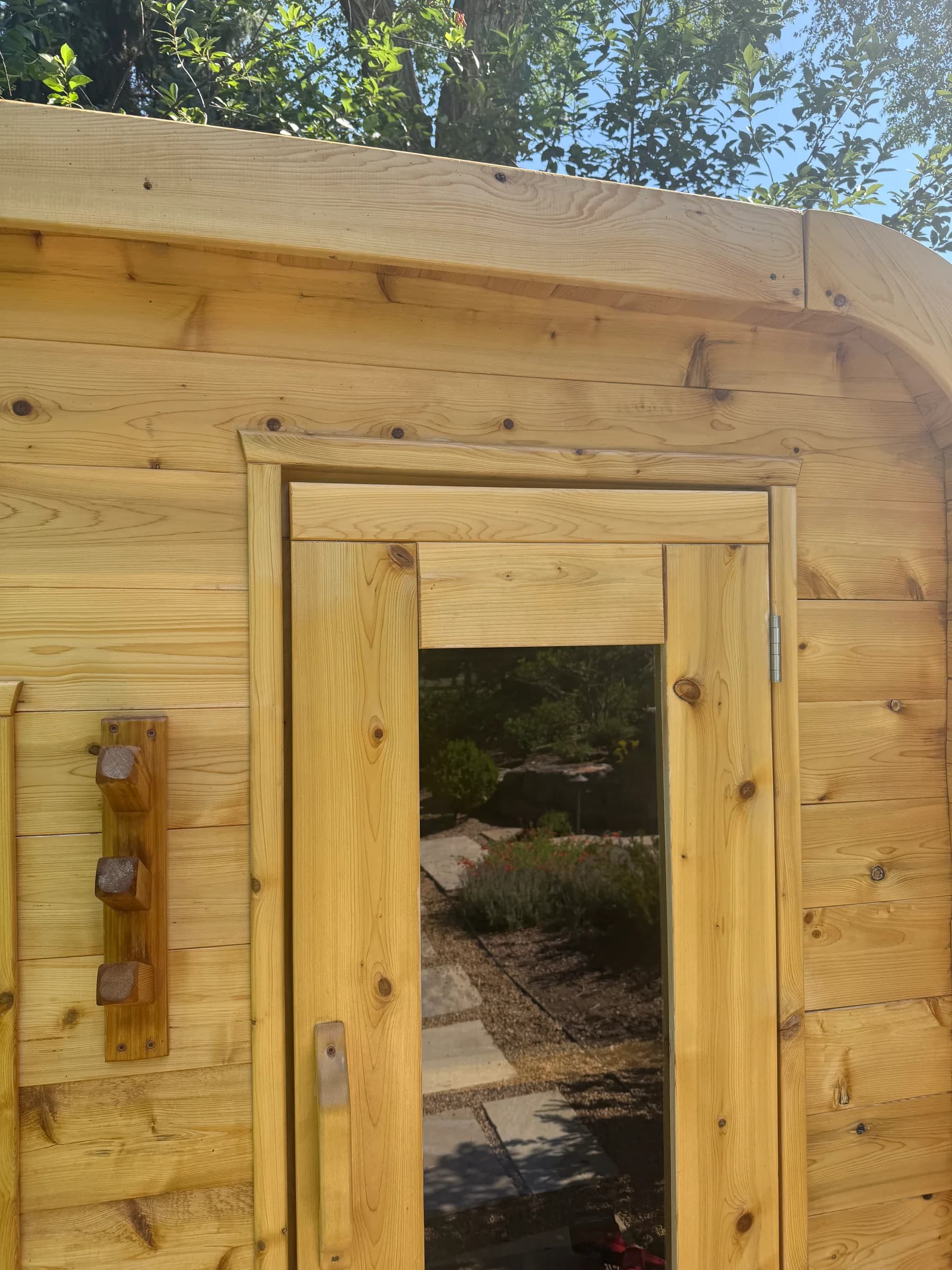 A close detail of a cedar sauna door and frame, a garden path reflected in its glass.