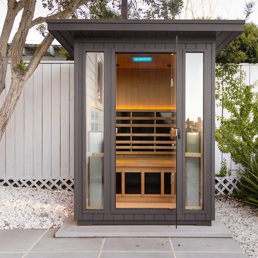 Clearlight's own image of a grey Sanctuary Outdoor 2 cabin, door open, standing on a raised concrete pad with white gravel on either side, square paving in front, and a white fence behind.