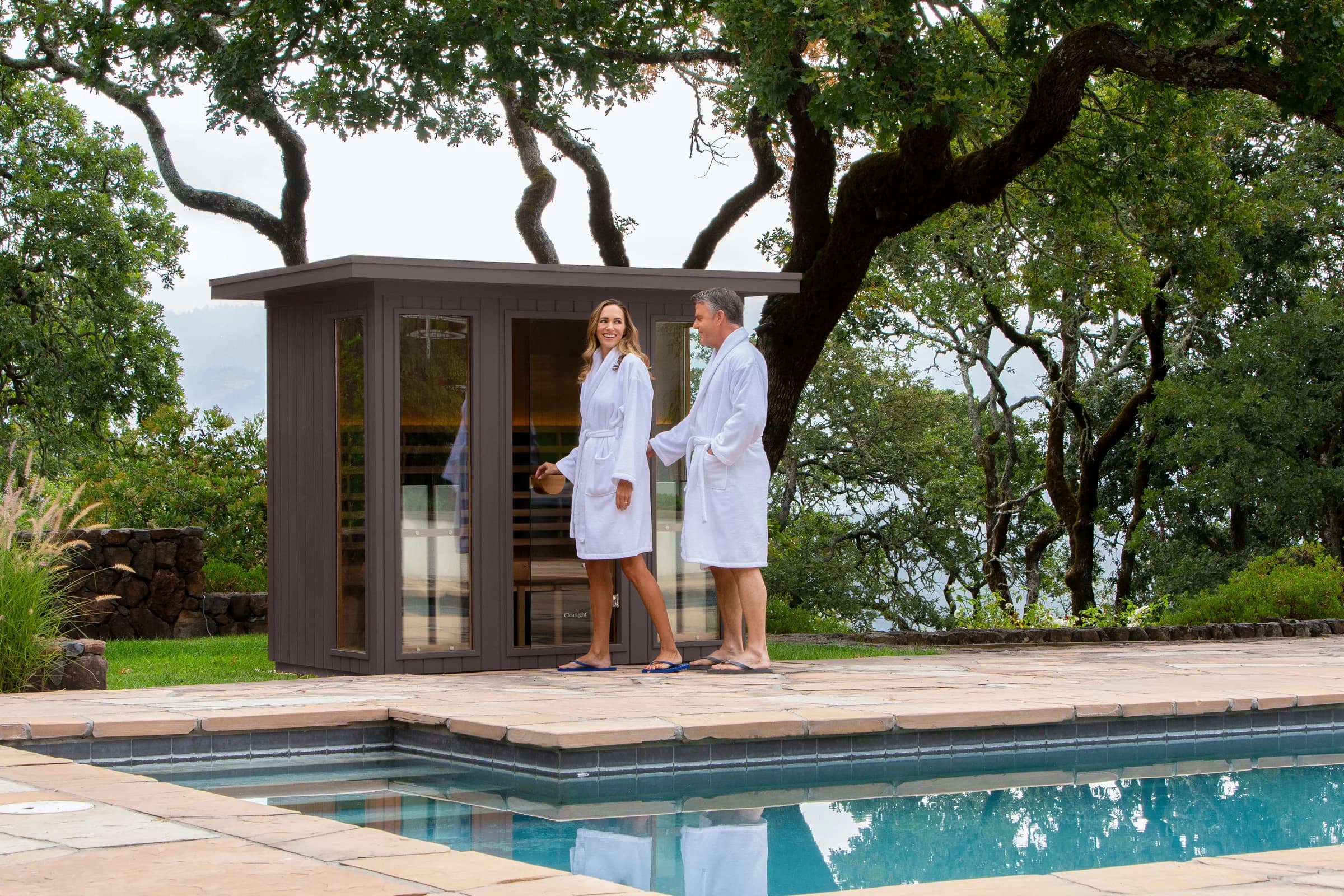 Clearlight's own image of a couple in white robes beside a grey Clearlight outdoor cabin on a flagstone pool deck under old oak trees.