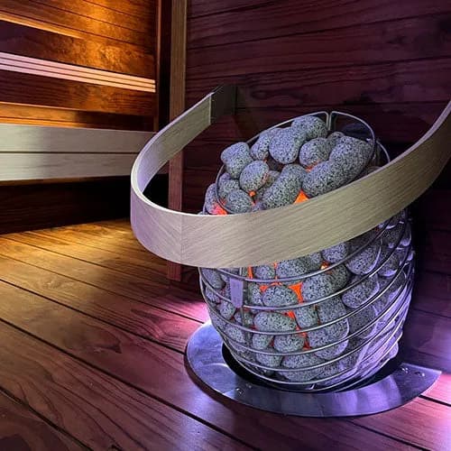 HUUM Drop heater glowing orange among speckled stones in a purple lit sauna corner with curved wood rail