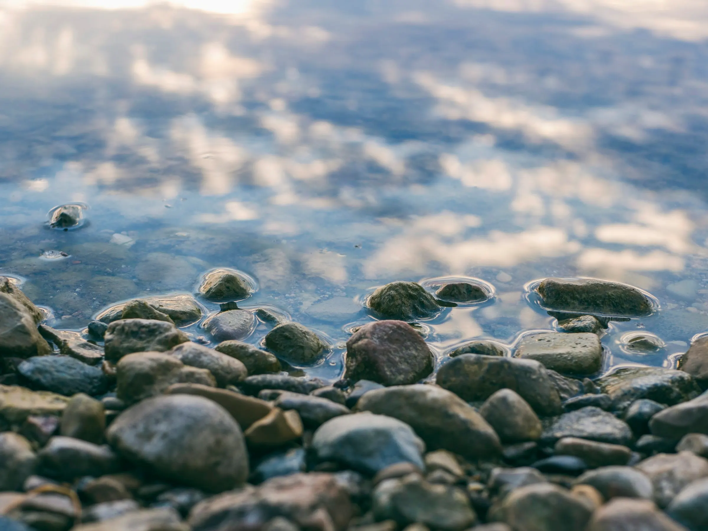 Rounded pebbles at the edge of a lake, with clouds reflected in the water.