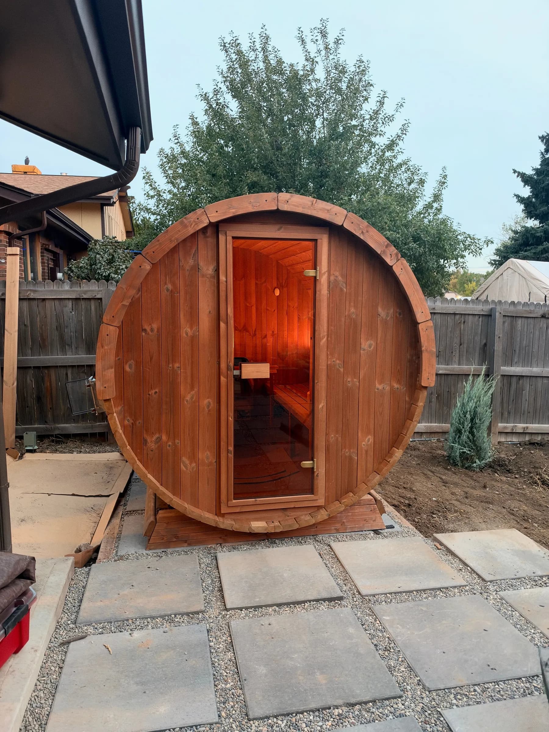 A finished cedar barrel sauna with a glass door, lit inside, at the end of a new path of square pavers set in gravel.