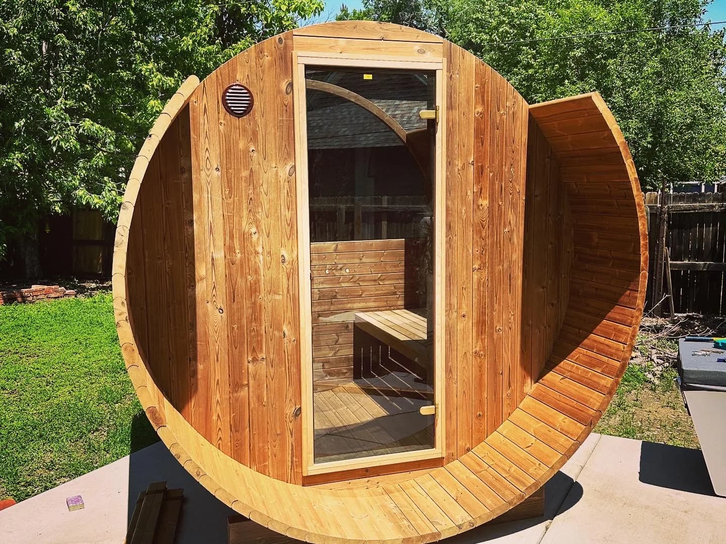 A cedar barrel sauna part way through assembly on a concrete pad in a fenced back yard: the glass-door end wall standing and the staves up both sides, the top of the shell still open to the sky, loose staves on the pad.