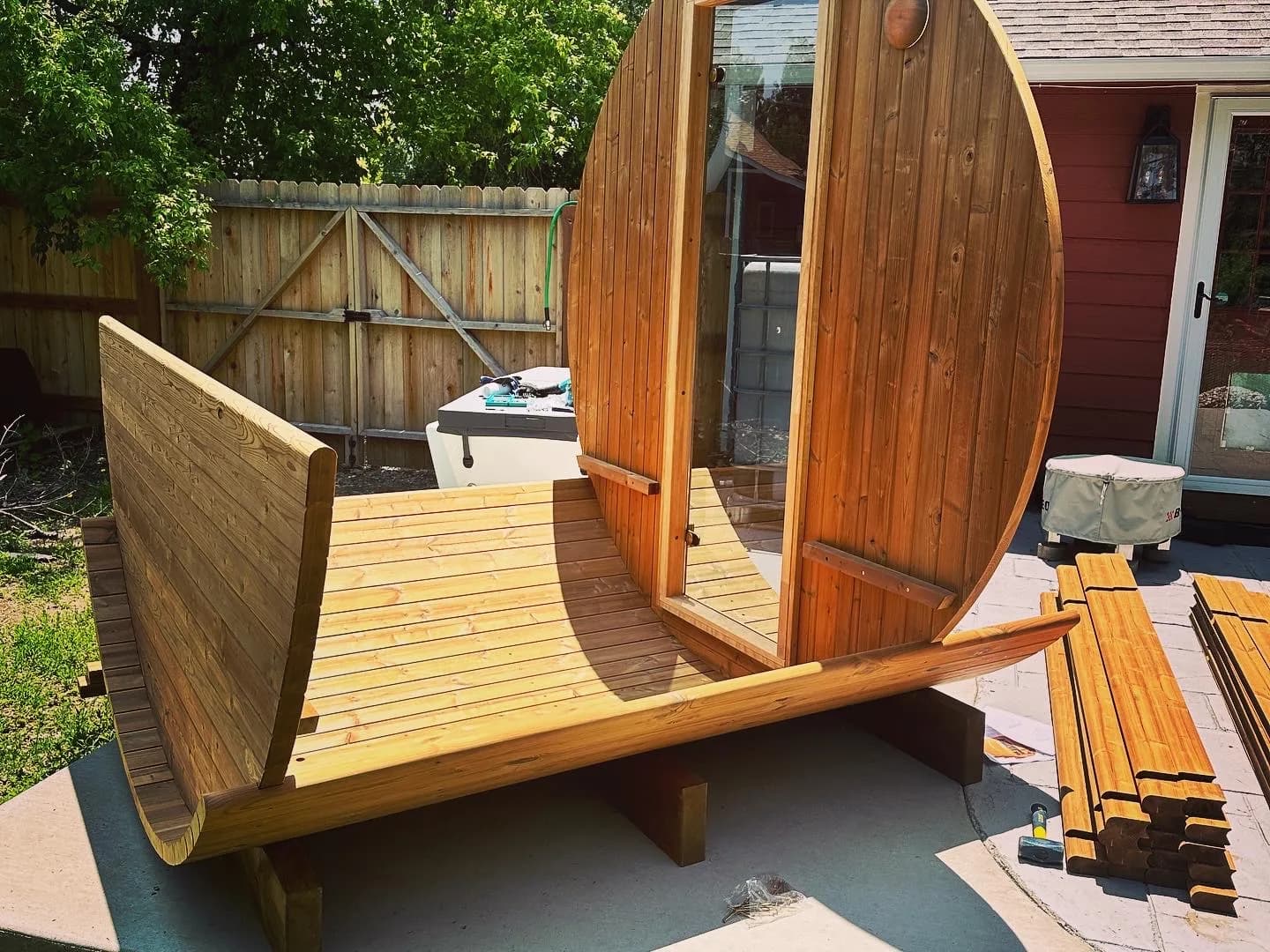A barrel sauna part way through assembly on its cradles, the floor laid and the front wall standing, with the remaining staves stacked on the patio alongside.