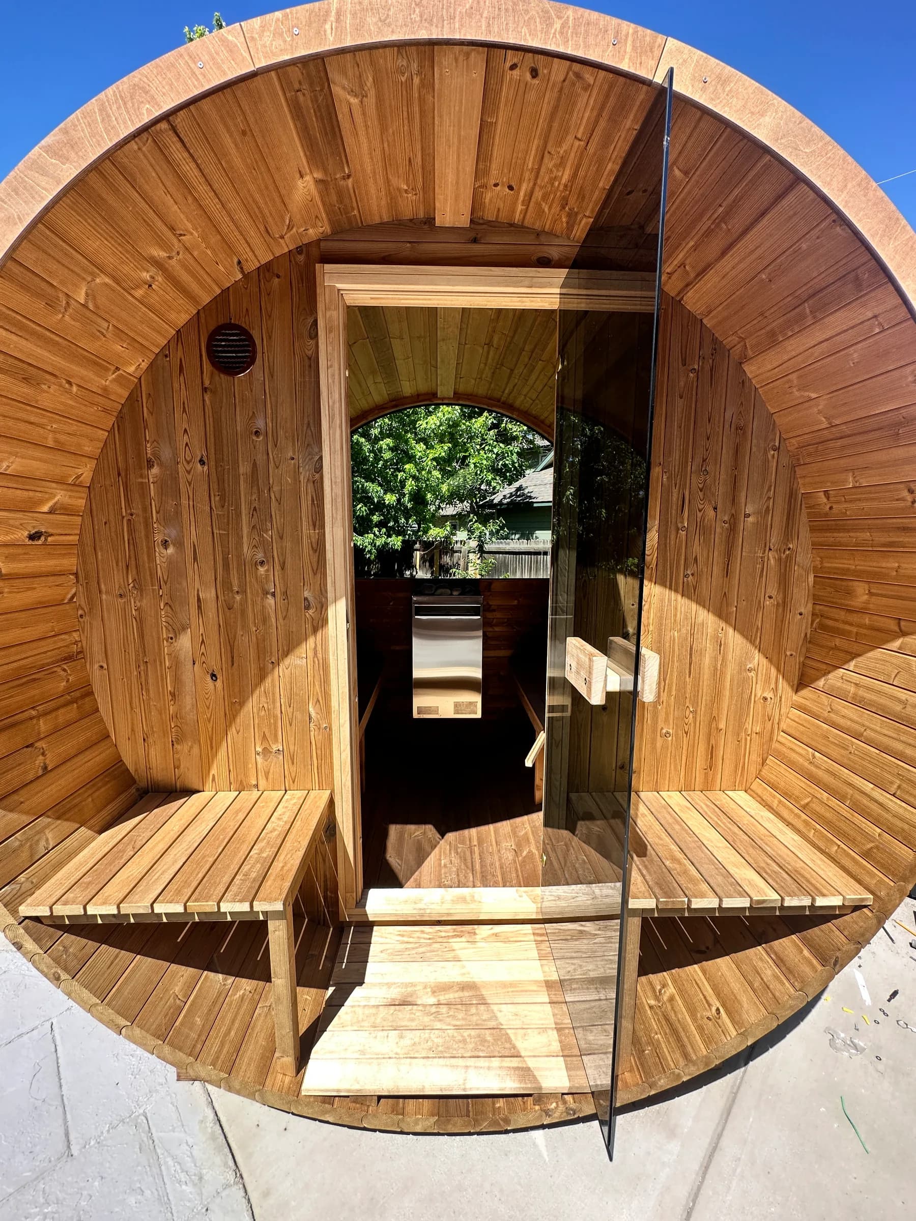 A cedar barrel sauna with its glass door open, benches on the front porch, and the interior visible through to a rear window onto trees.