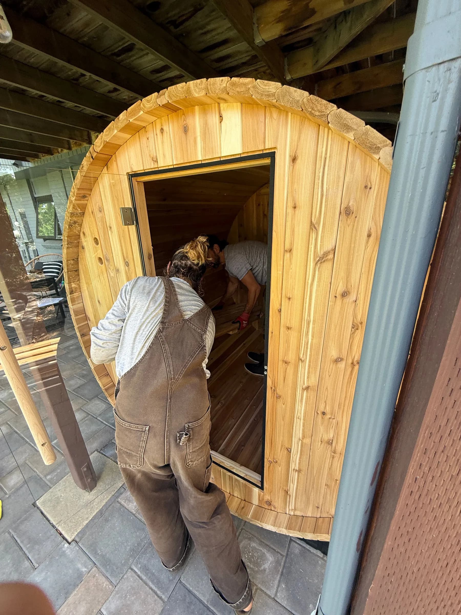 An installer in overalls stepping into the open door of a pale wood barrel sauna under a covered patio.