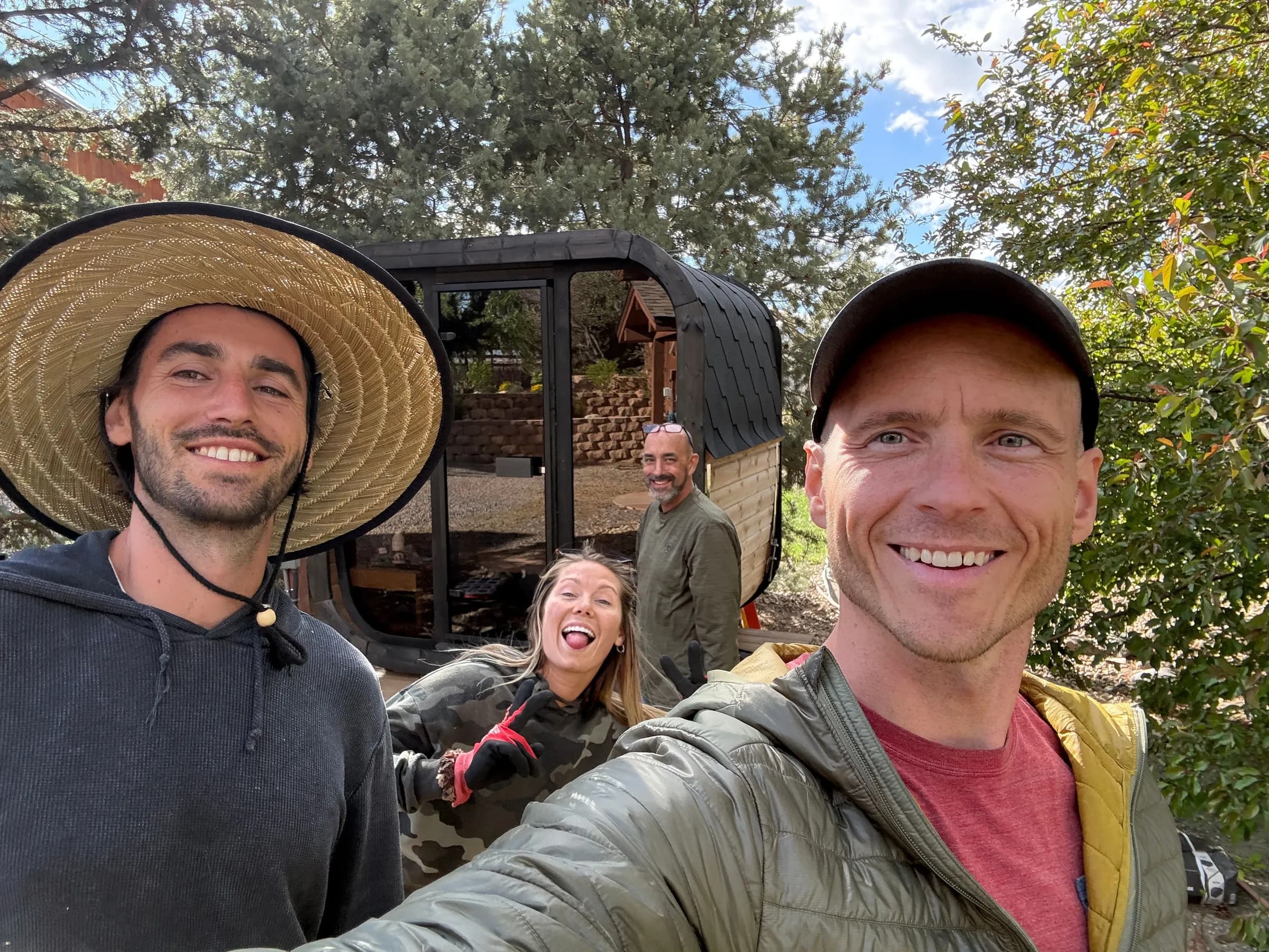 Four of our crew smiling on site in front of a dark cube sauna they are building, trees behind.