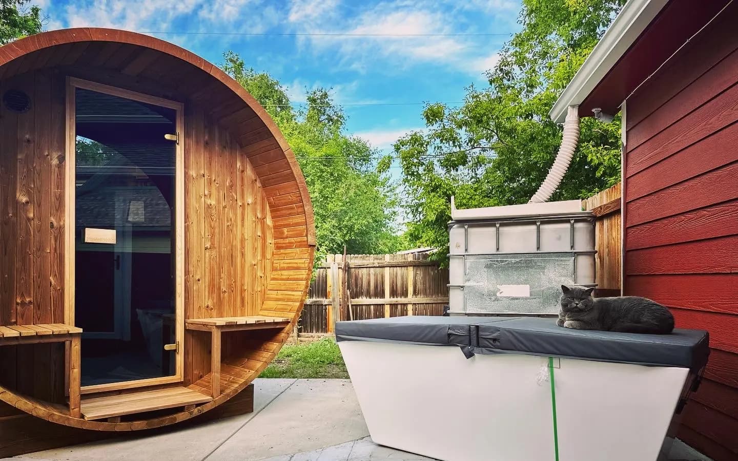 A cedar barrel sauna with a glass door beside a covered cold plunge tub on a stone patio, a grey cat asleep on the tub cover.
