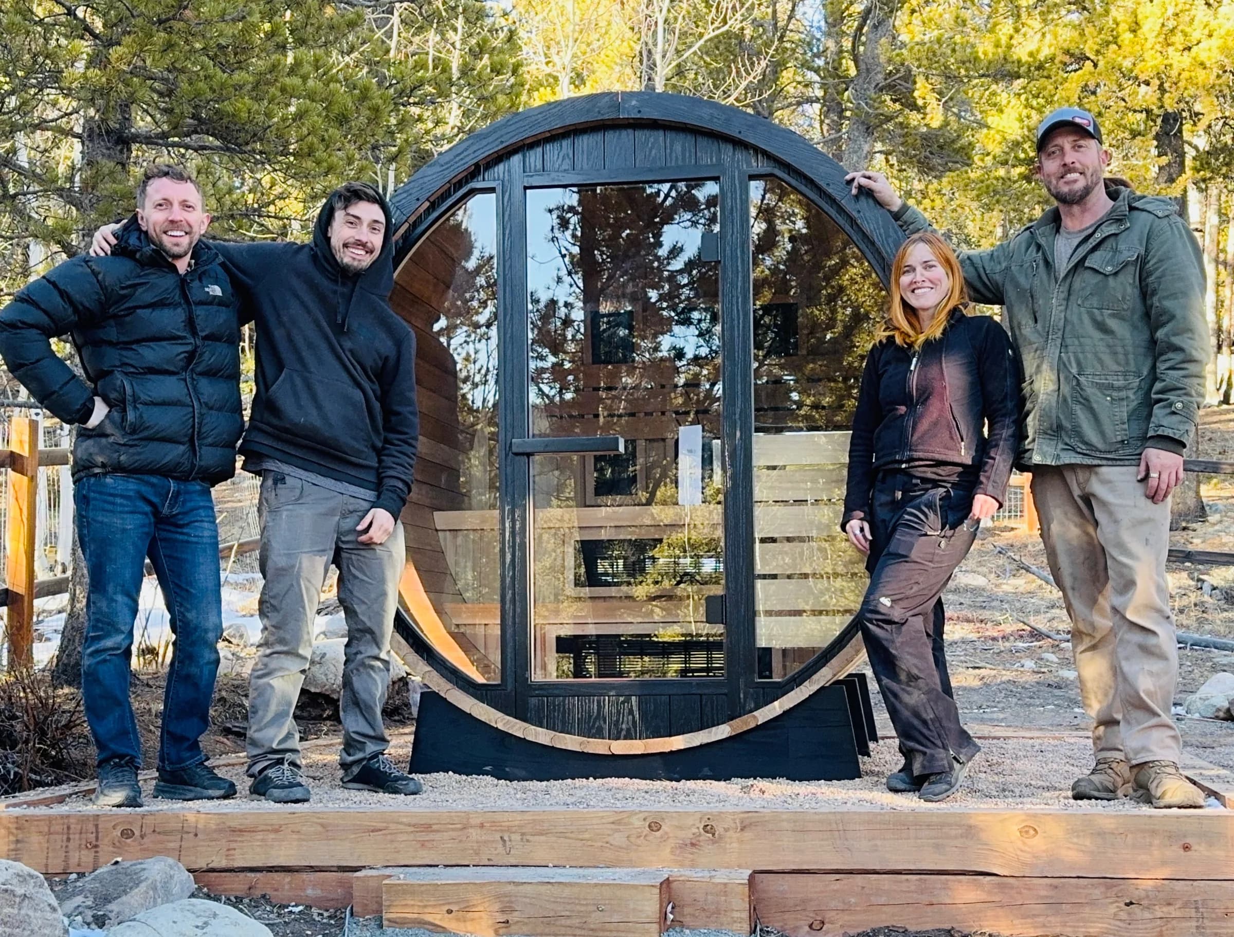 Four members of our crew standing either side of a finished black barrel sauna with a round glass front, on a timber-framed gravel base among pines and aspens.