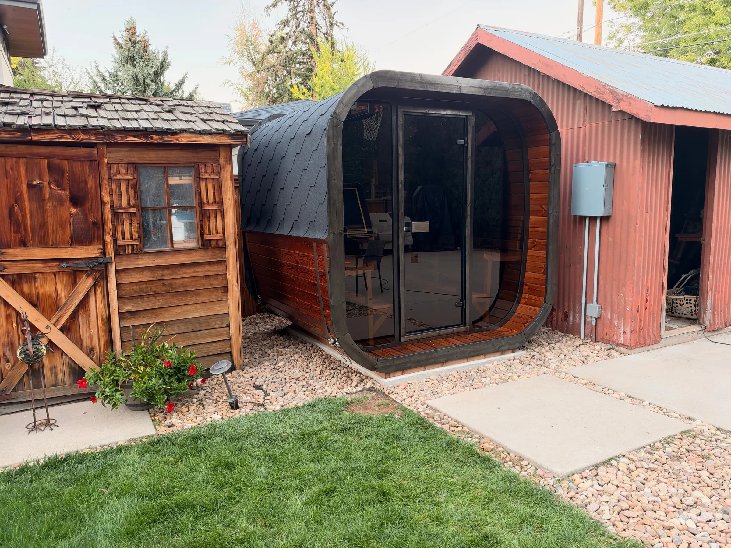 A finished cube sauna with a dark rounded shell and glass doors, set between a timber shed and a red barn-style shed on a gravel and paver base.