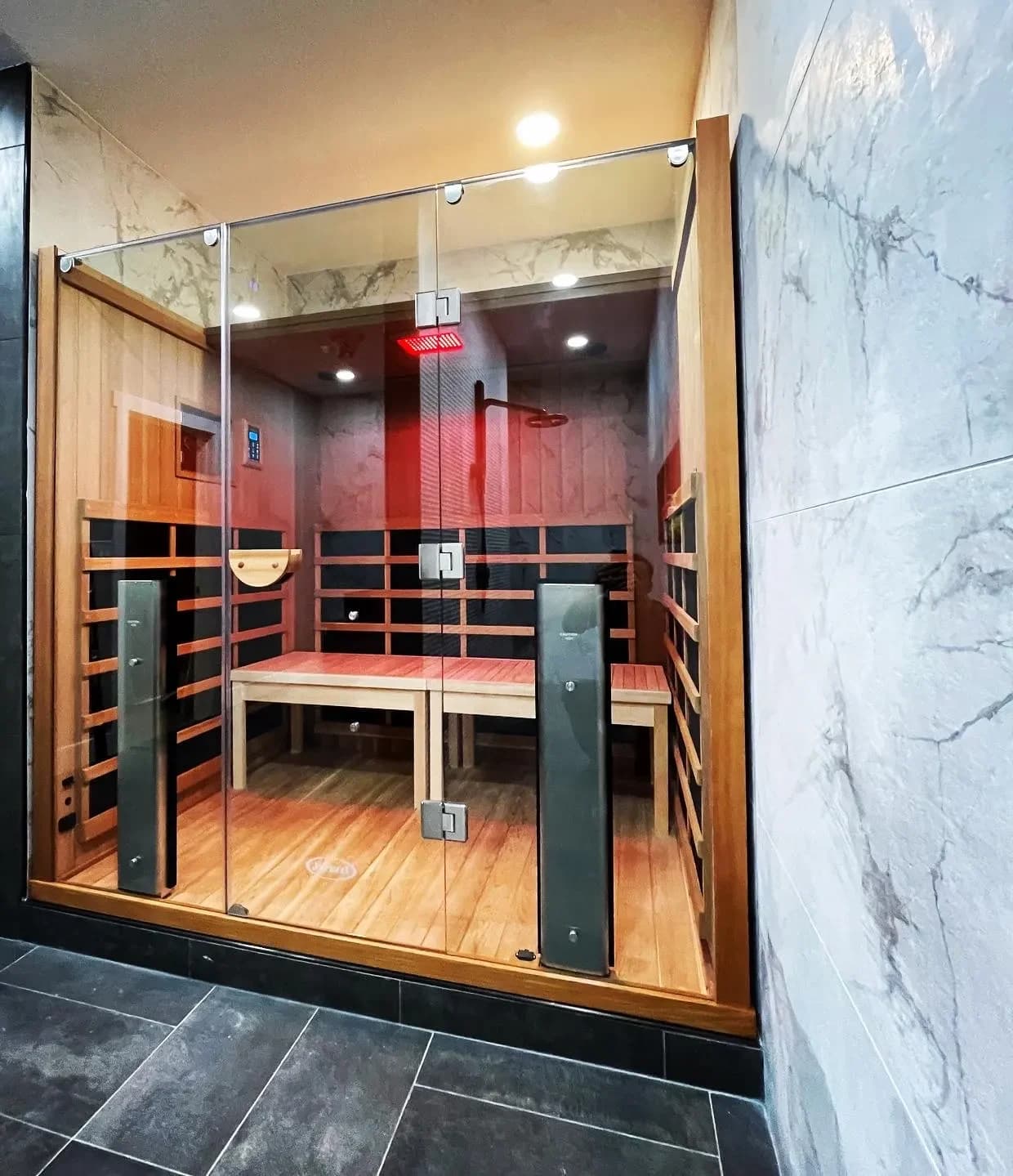 A glass-fronted indoor sauna with two benches and a lit ceiling, set into a marble-tiled room with a dark slate floor.
