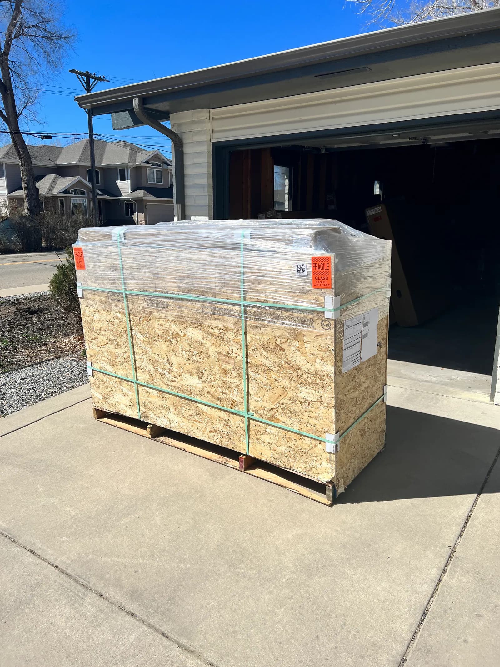 A crated sauna, shrink wrapped and banded to a pallet, standing on a driveway outside an open garage.