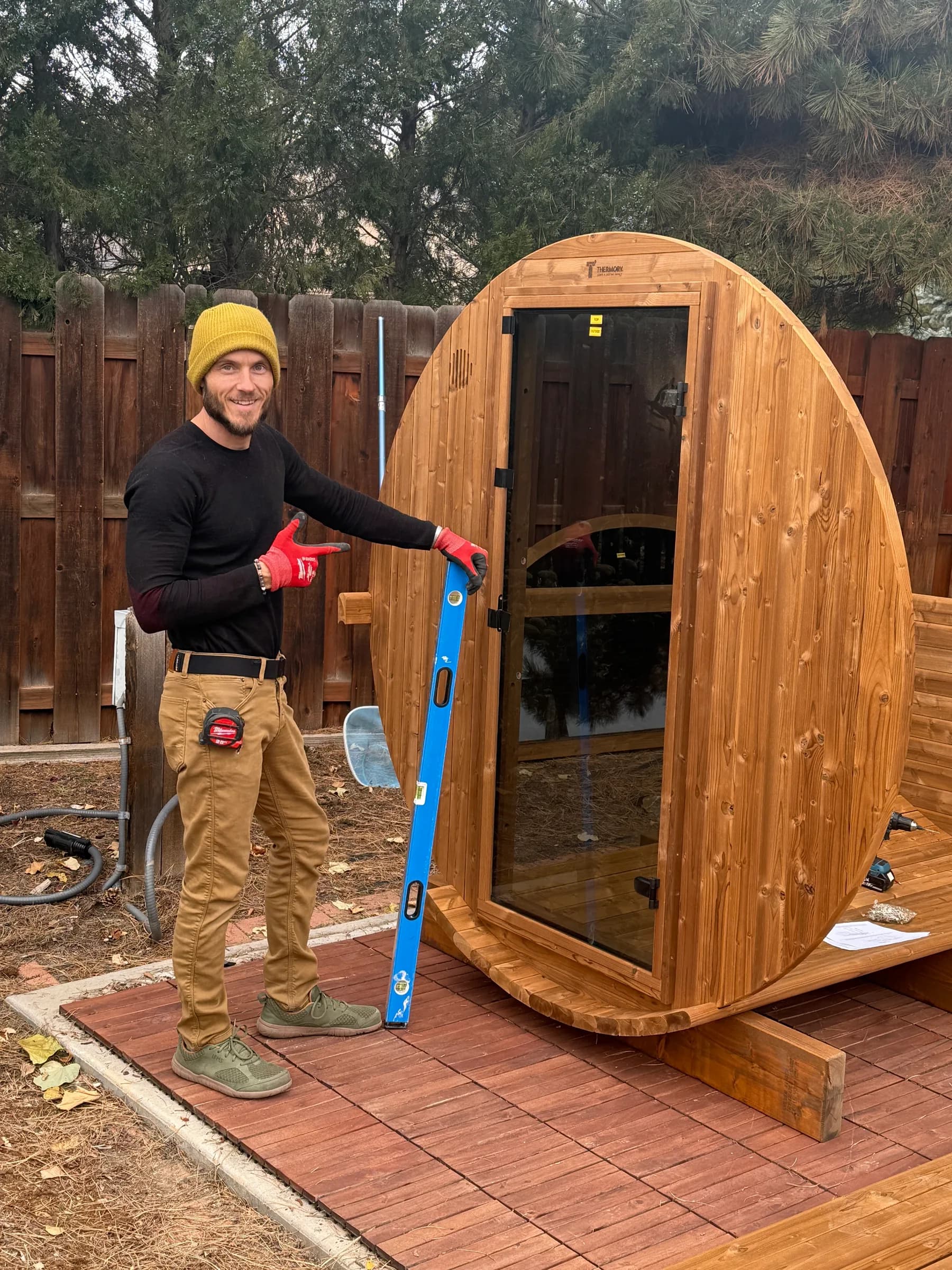 An installer in a yellow beanie pointing to an upright level beside the glass-door end wall of a barrel sauna going up on a patio of red-stained deck tiles, a drill and the instructions on the floor boards behind it.