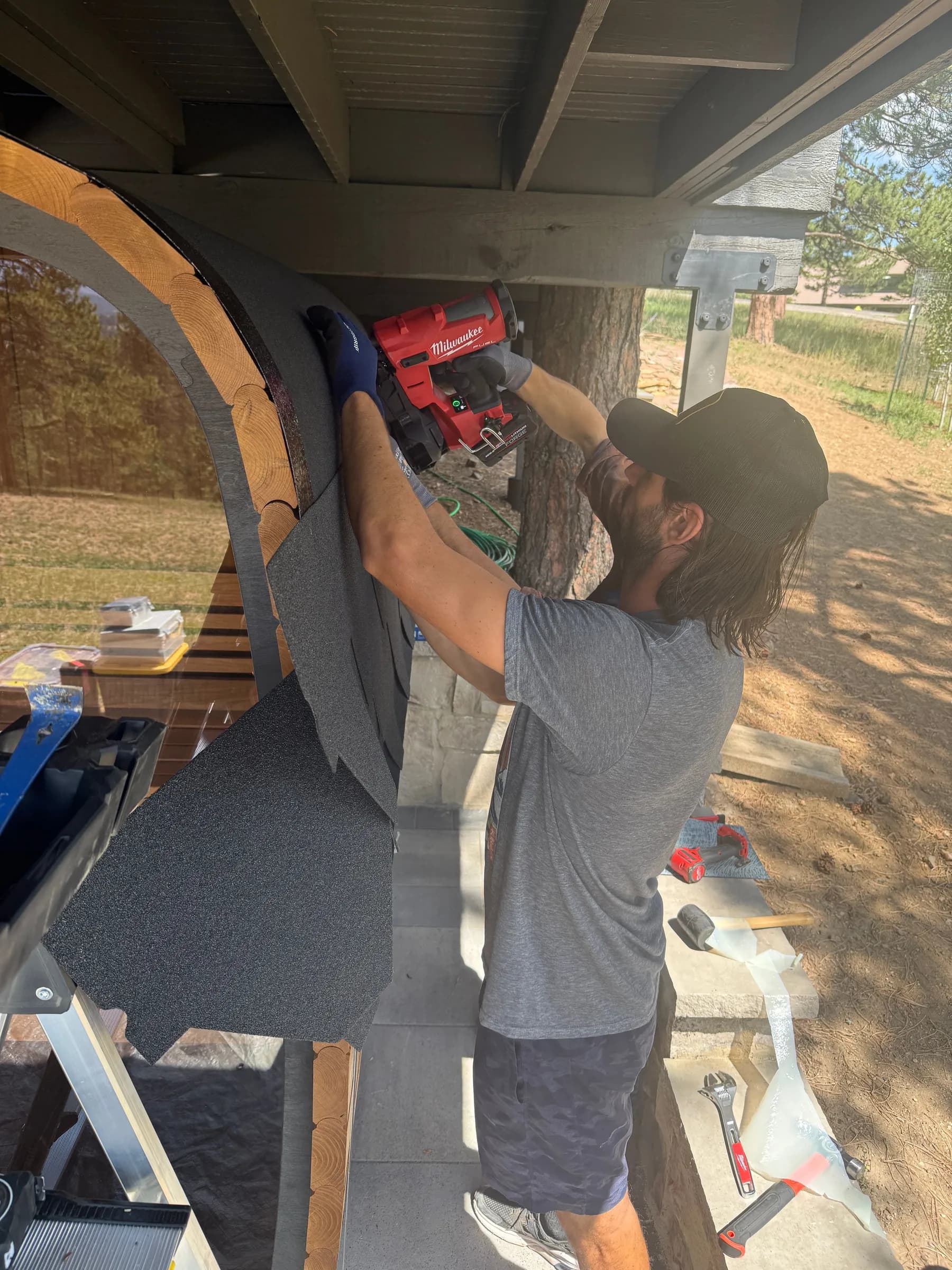 An installer fixing the dark shell of a cube sauna with a drill under a covered patio, the sauna's rounded front beside him.