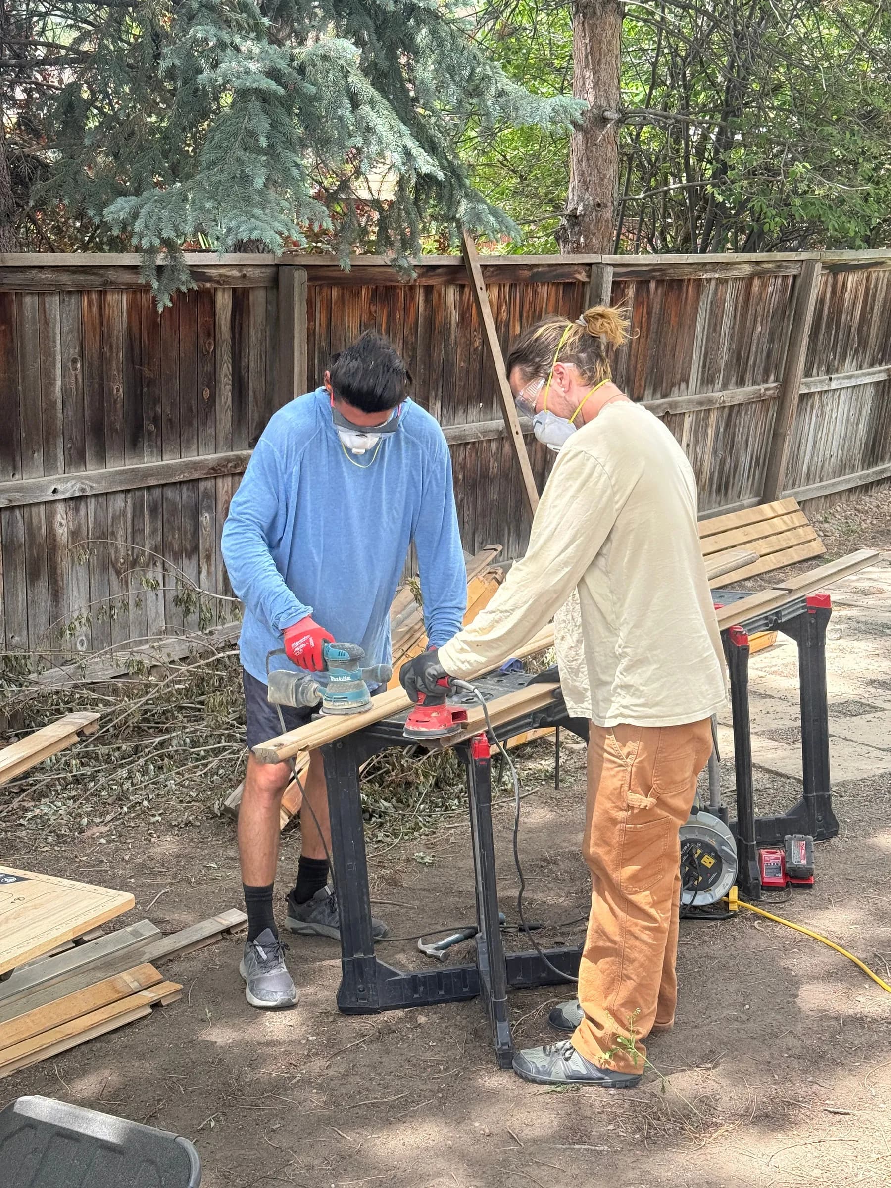 Two of our crew in dust masks cutting boards on a saw stand in a back yard, timber stacked by the fence.