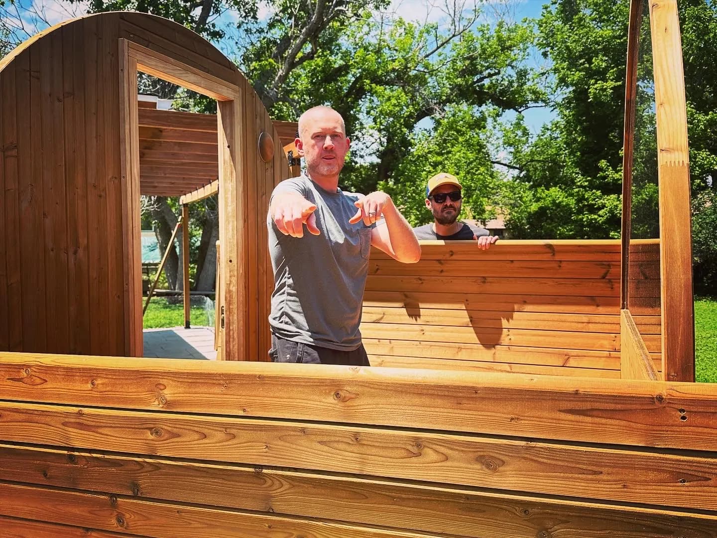 A barrel sauna half built in a sunny backyard, one man standing inside the waist-high walls and pointing toward the camera while a second, in a cap and sunglasses, looks over the far wall.