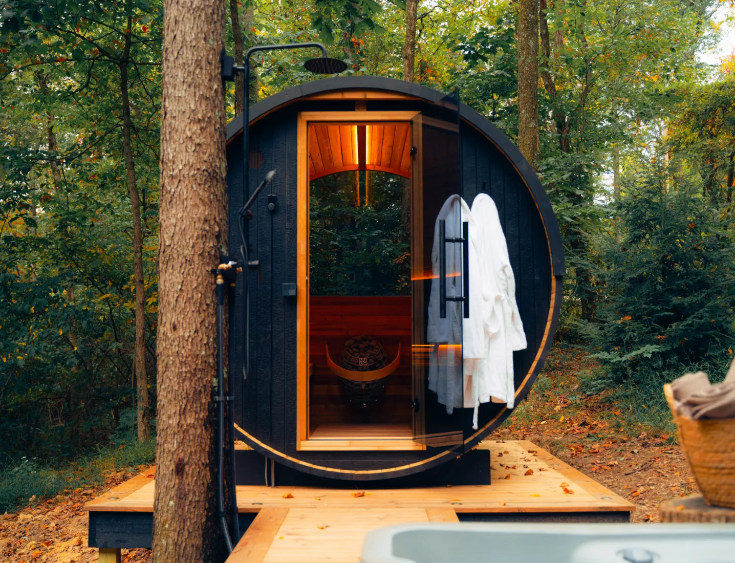 Thermory Ignite barrel sauna with round door open in a forest setting with bath robes and outdoor shower — Sauna Friend Sanctuary Supply
